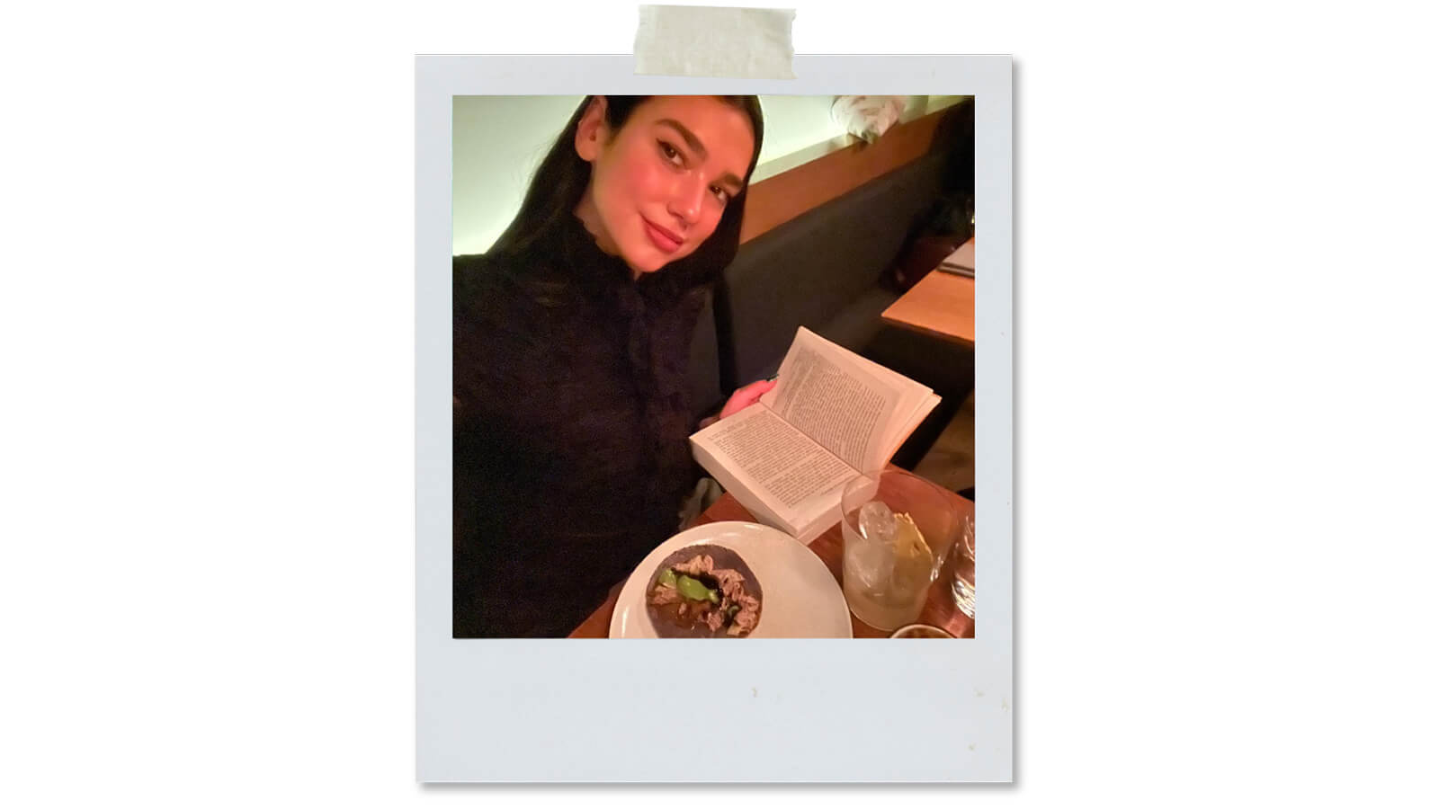 Dua Lipa reads book at dinner table, where she is eating a taco