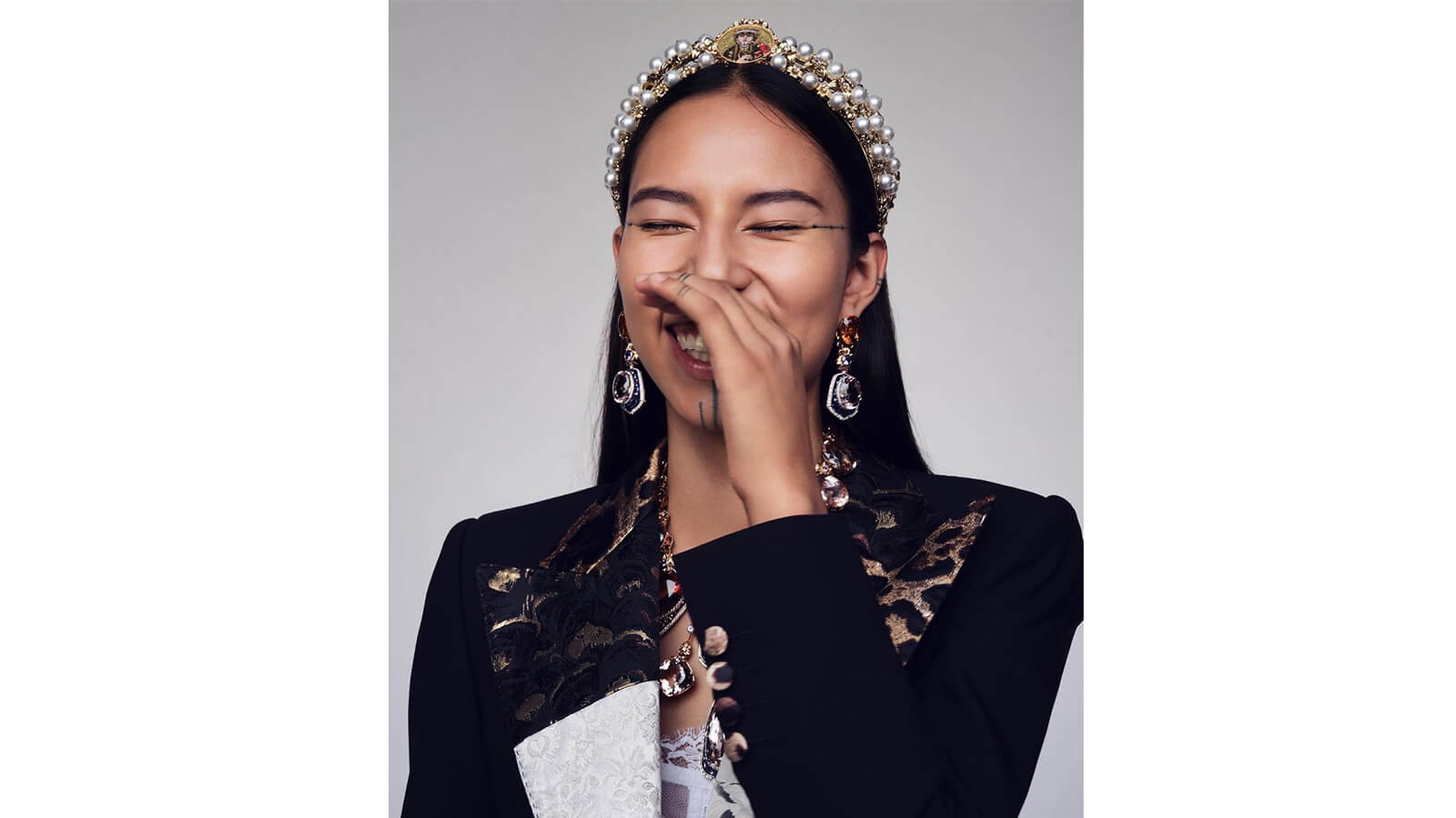 Brunette model with tribal facial tattoos, laughs into hand wearing pearl crown