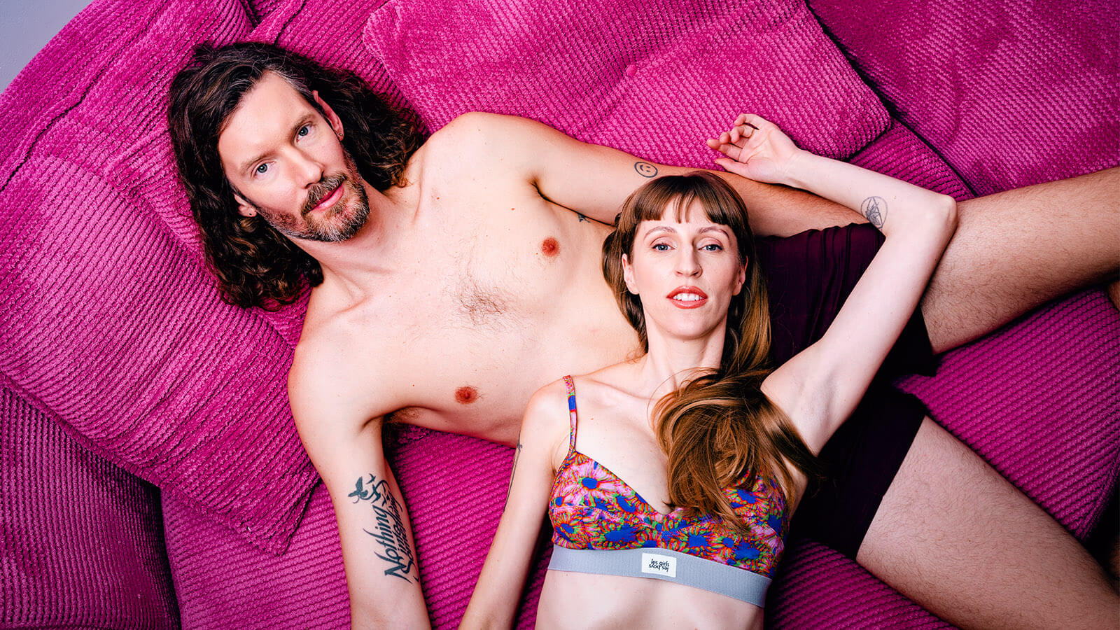 Image of couple, sex and relationship coaches and creators of the Lacey and Flynn Have Sex podcast on pink sofa in underwear