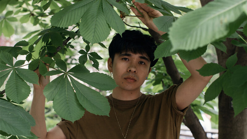 Portrait of writer and poet Ocean Vuong standing under the branches of a tree