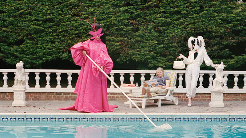 Photograph of a model cleaning a pool wearing a full-length pink gown and pink Schiaparelli haute couture mask by Stephen Jones, a model wearing a dog mask and an older woman in casual clothes reading a magazine on a sun lounger