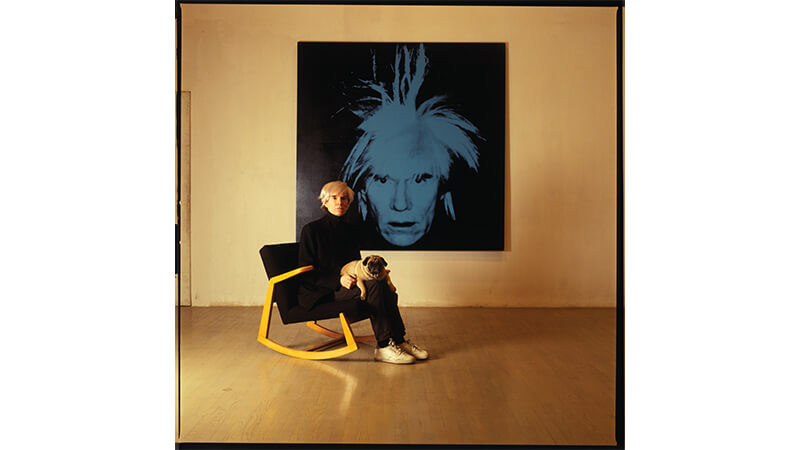 Portrait of artist Andy Warhol, with dog, sitting in front of painting titled Self Portrait, 1986